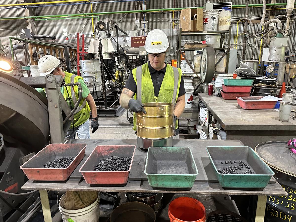 Man in hard hat and safety vest stands at table holding four trays of iron pellets.
