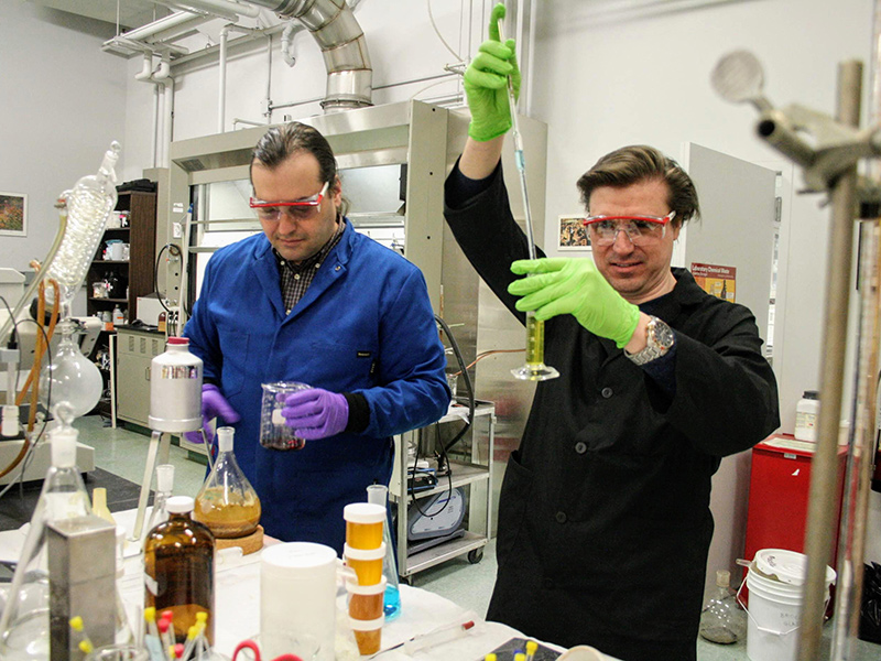 Two scientists wearing lab coats and safety glasses pipette liquid into beakers