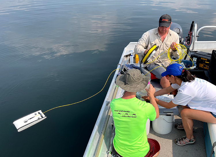 Three people use water monitoring equipment from small flat bottomed boat