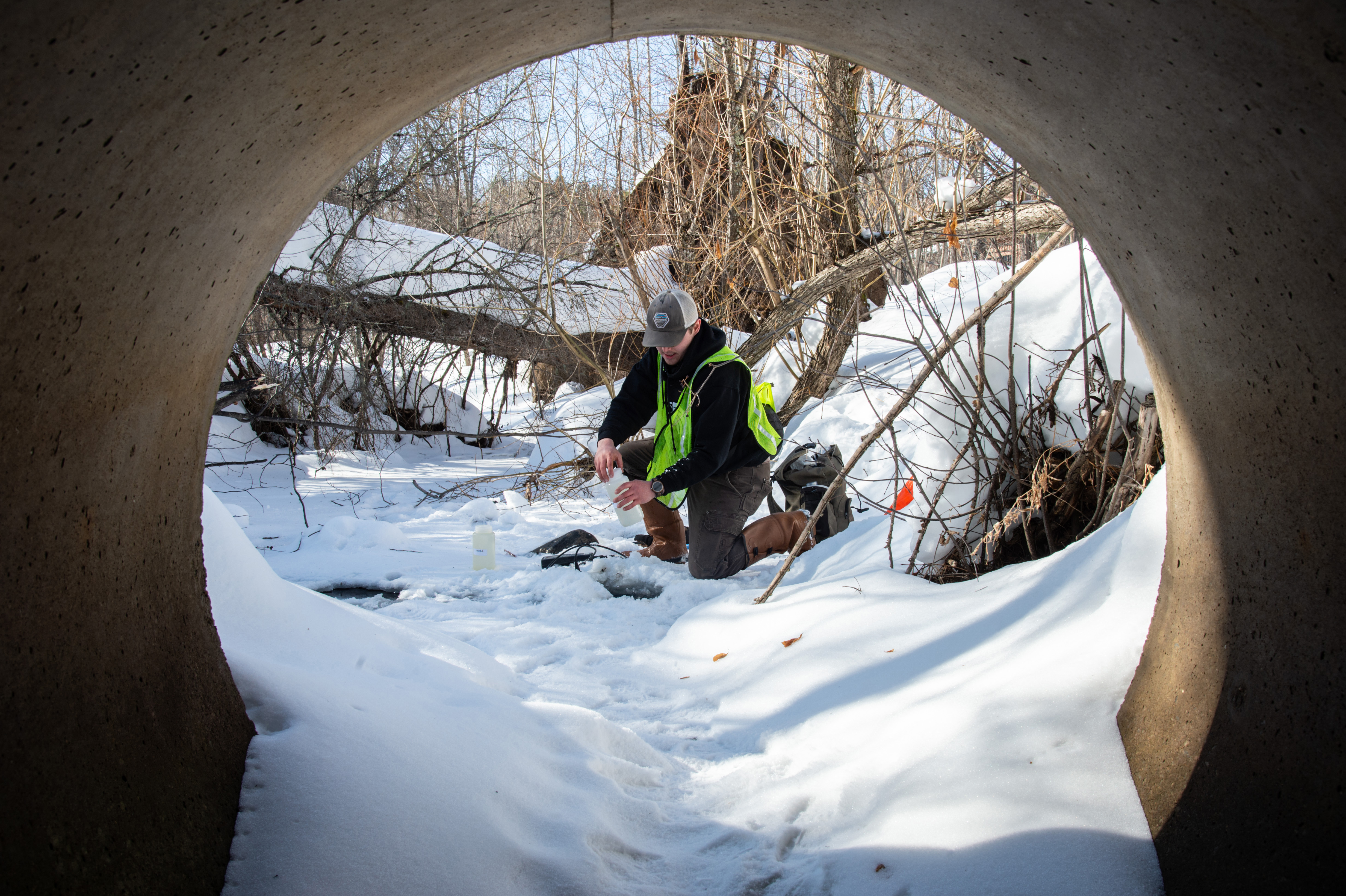 Man kneeling on frozen stream collecting water through hole in ice.