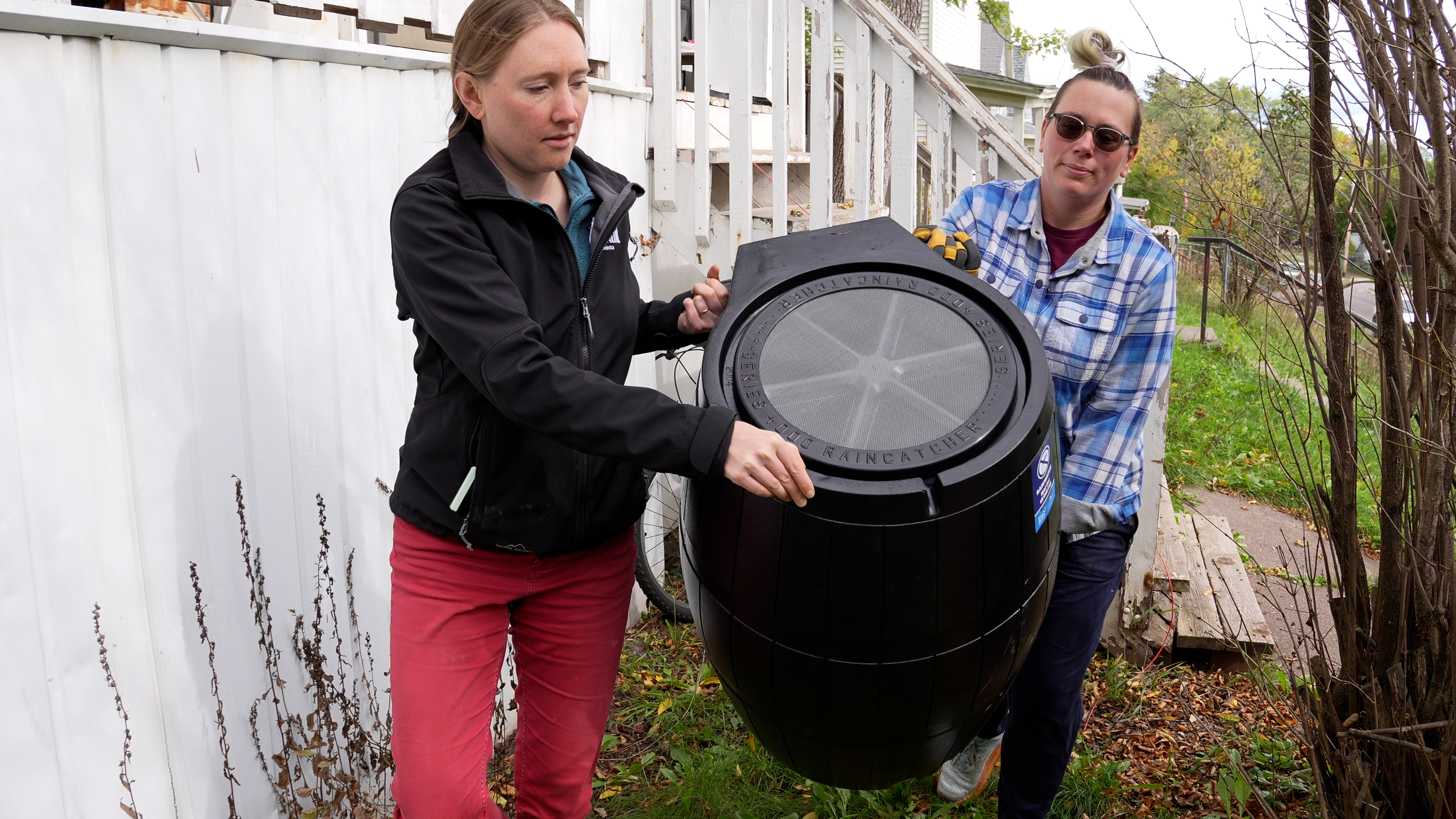 Two women carry a rain barrel alongside a residence.
