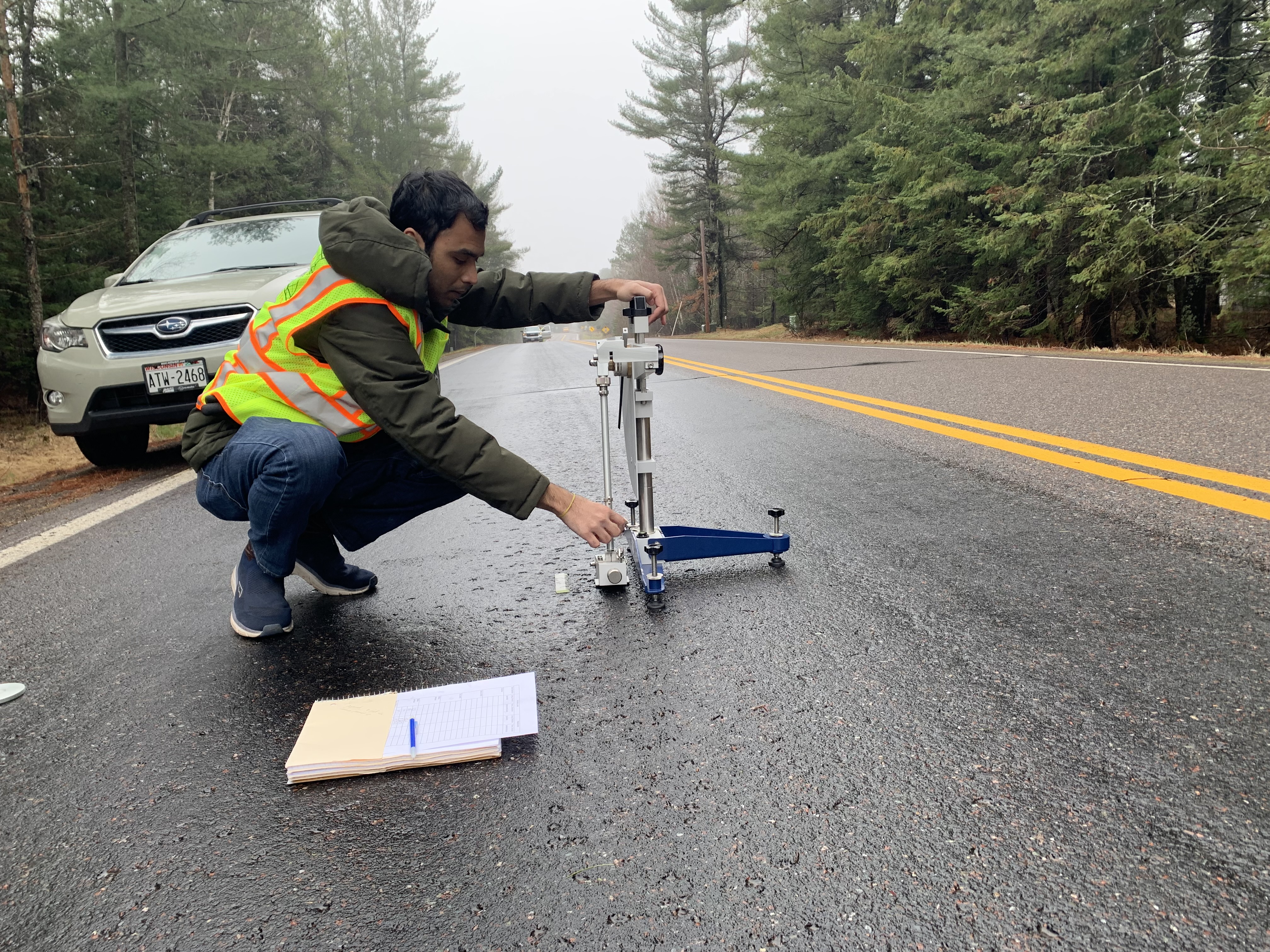 A man in a safety vest crouches on a road holding up a testing apparatus.