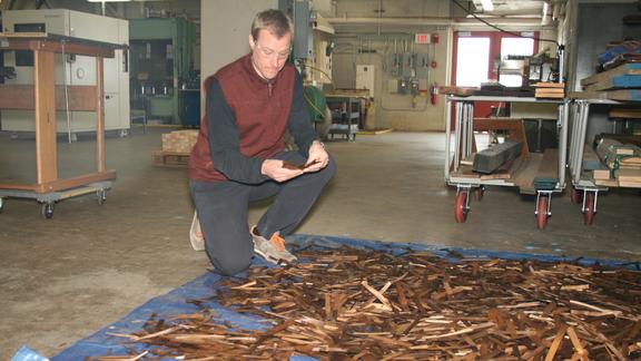 A man kneels in front of a tarp on the floor covered with dark wood shavings.