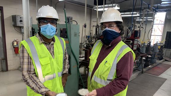 Two men wearing safety PPE holding small dish each with powdery material.