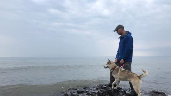 A man and large dog stand on a rock on shore of very large lake.