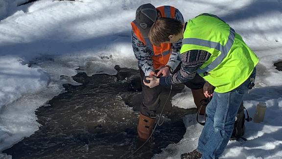 Two men wearing safety vests stand on frozen stream bent over hole in ice.