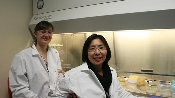 Two women in white lab coats and safety glasses by a large machine.