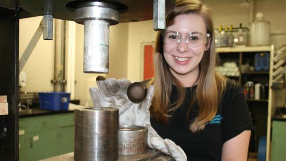 Woman is framed by machinery, holding a small brown disk