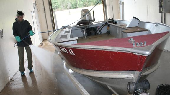 A man with face guard, gloves and rain coat operates car wash wand on boat