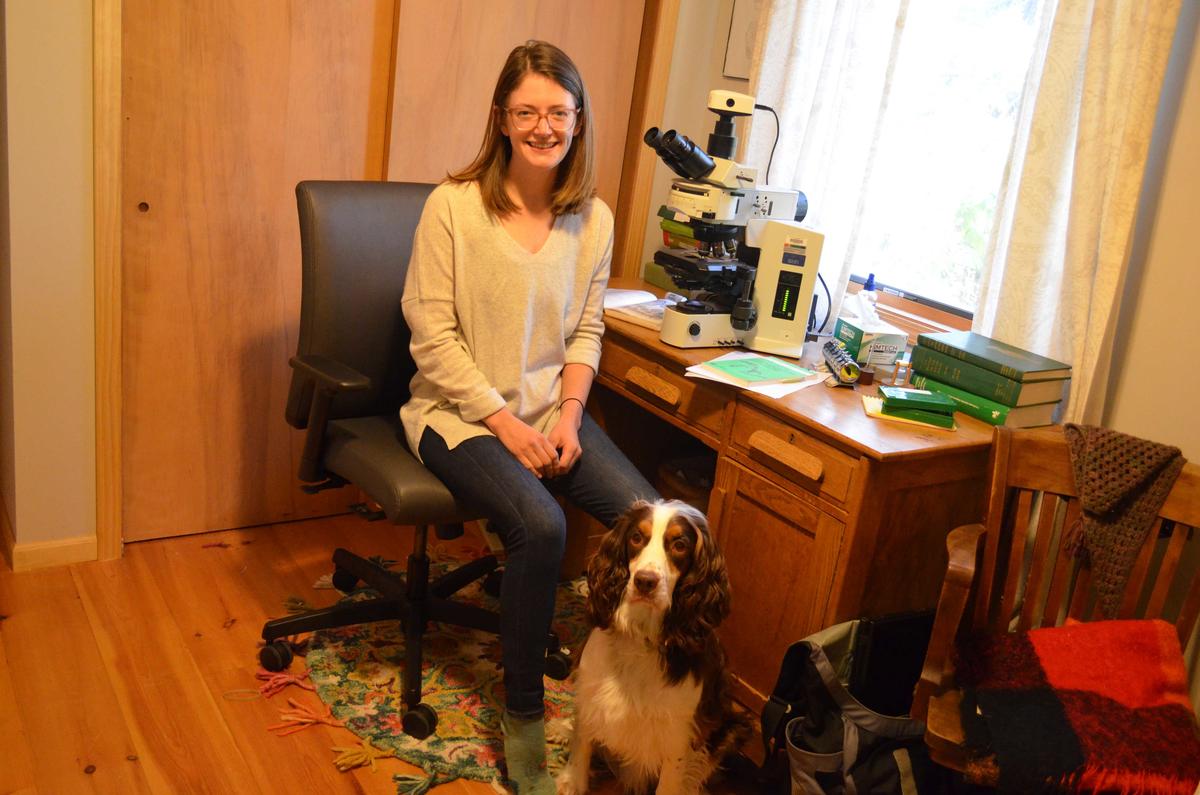 Woman sits a desk with microscope on it; dog at her feet.