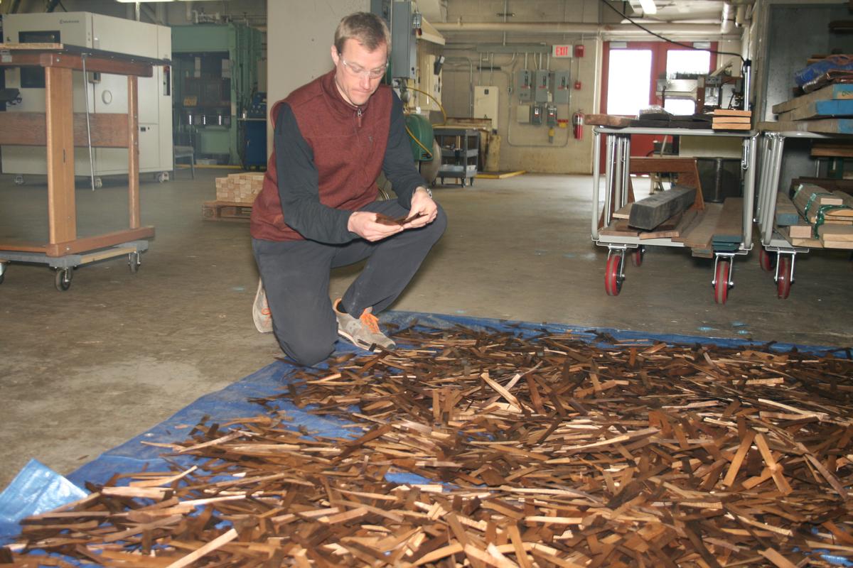 A man kneels in front of a tarp on the floor covered with dark wood shavings.