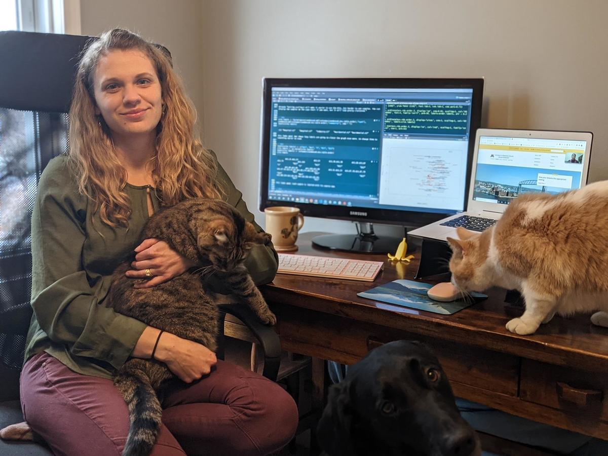 A woman sits at a home office desk with dog and two cats
