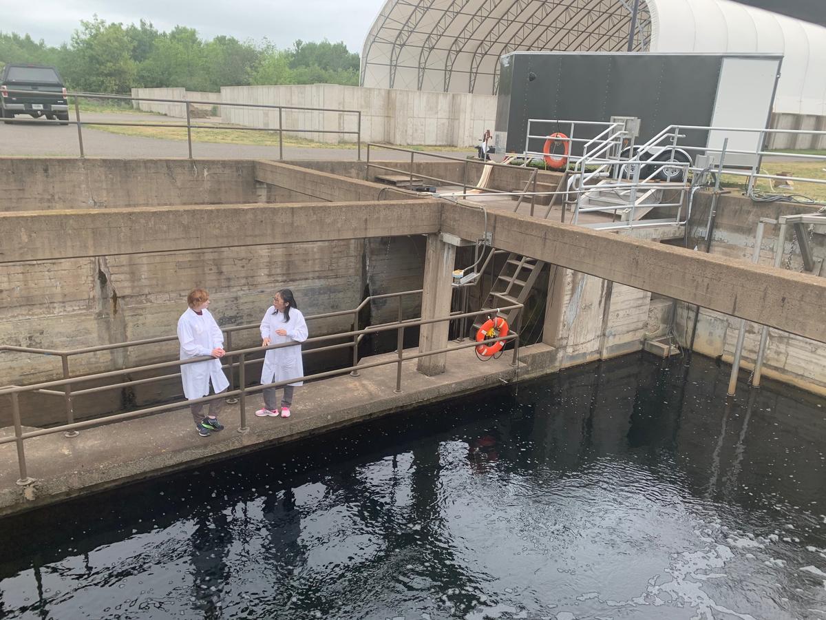Two scientists wearing lab coats stand on cement walkway next to water resevoir