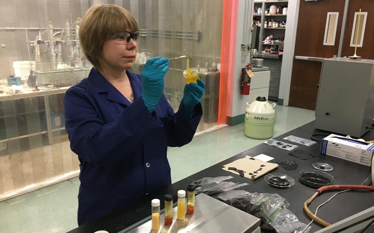 Woman in lab pulls thick yellow substance from small jar with glass rod