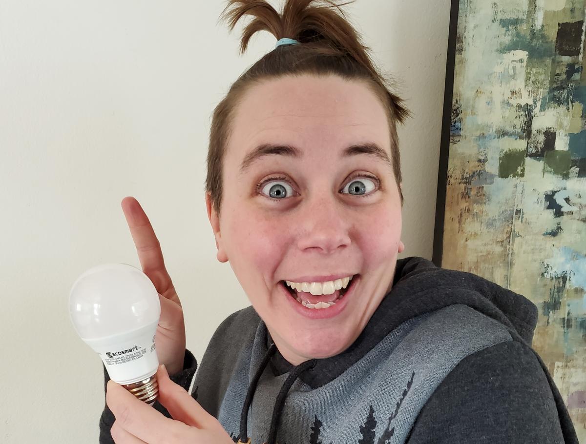 Woman holds light bulb to left side of face pointing to lightbulb, smiling.