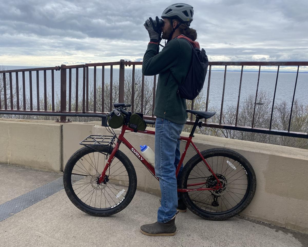 Man stands next to bicycle while looking through binoculars.