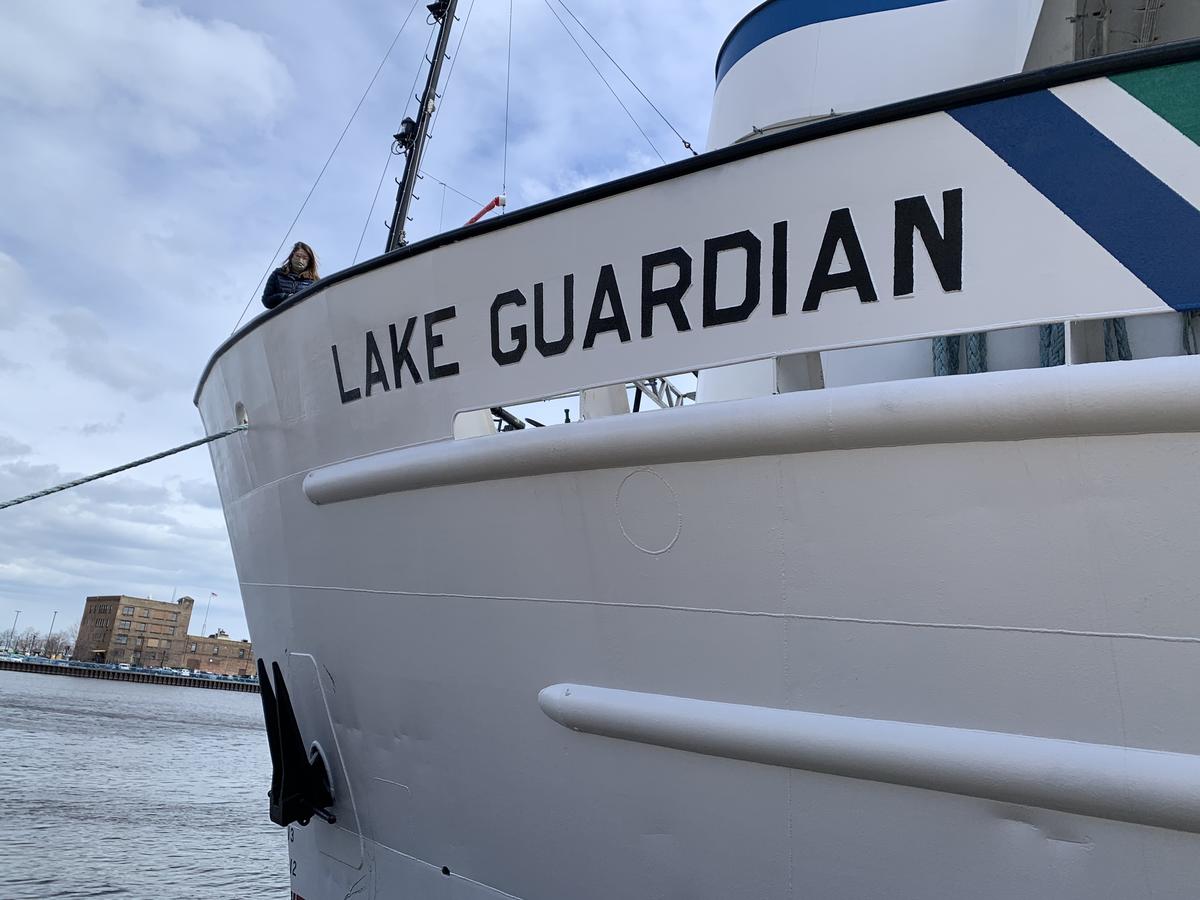 Woman stands at far end of large boat with Lake Guardian printed in large letters on the bow.