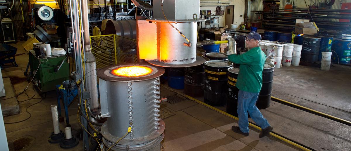 A man closes metal lid over bright hot upright kiln in industrial setting.