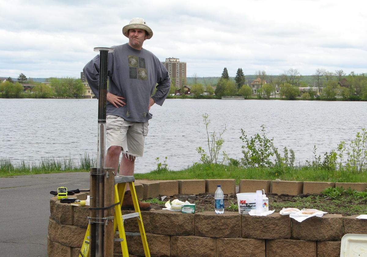 A man stands on a short ladder with lake in background looking at long verticle tube