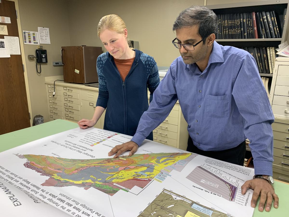 A woman and man stand behind a desk looking at a large map.