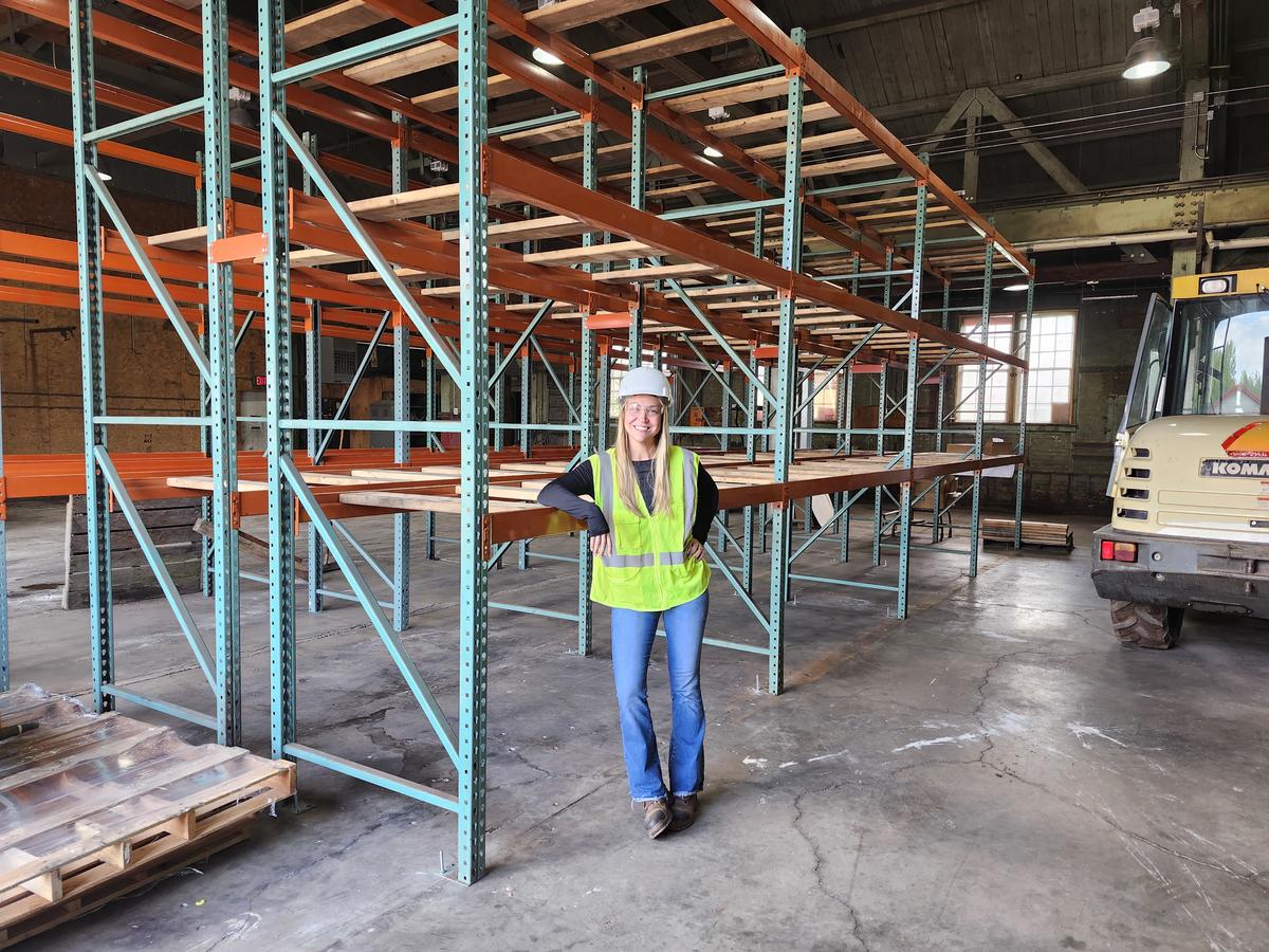 A woman wearing PPE stands next to industrial shelving in warehouse.