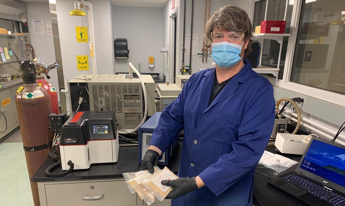 Man in blue lab coat in lab setting holds four clear packets containing wood chips