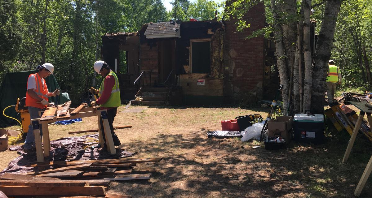 Two men in construction gear work outside a partially torn apart house structure.