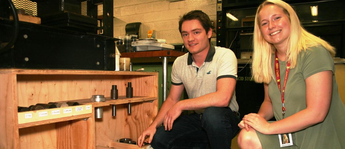 A male and female are kneeling next to wooden shelving in an industrial setting