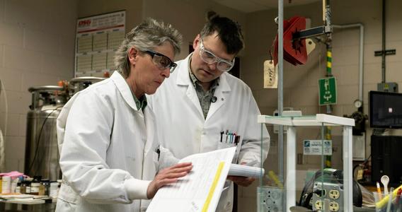 A woman and man wearing white lab coats look over a file in a lab setting.