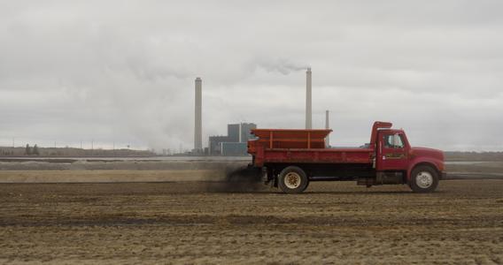 A truck pulling a spreader that is distributing dusty material on ground.