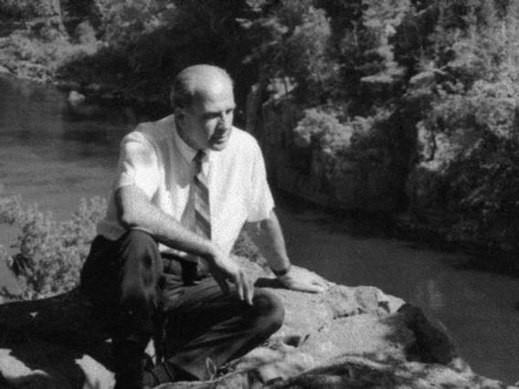 Man in white shirt and tie sits on rock by a river.