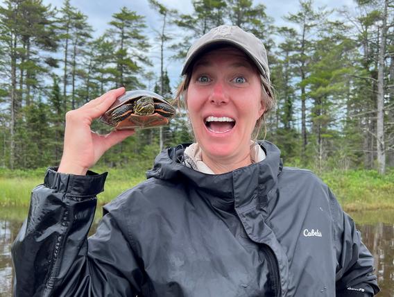 Woman smiles broadly while holding a wood turtle over her shoulder.