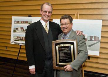 Two men pose for camera. Man on right holds a plaque.