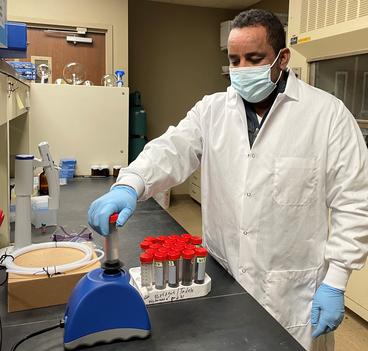 A man in white lab coat and face mask stands at a lab bench.