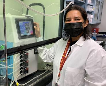 Woman in white lab coat and face mask stands by lab equipment