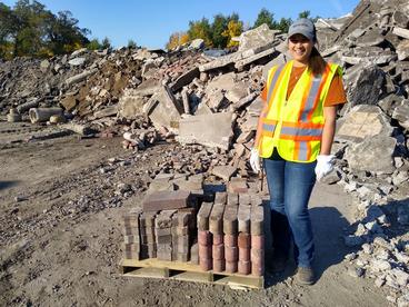 A woman stand next to a pallet of neatly stacked landscaping blocks, with a huge rubble pile in the background.