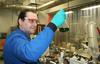 Man in blue lab coat and safety glasses looks at a vial with dark material inside.