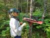 A woman wearing a head net looks at a small mammal trap attached to a tree.